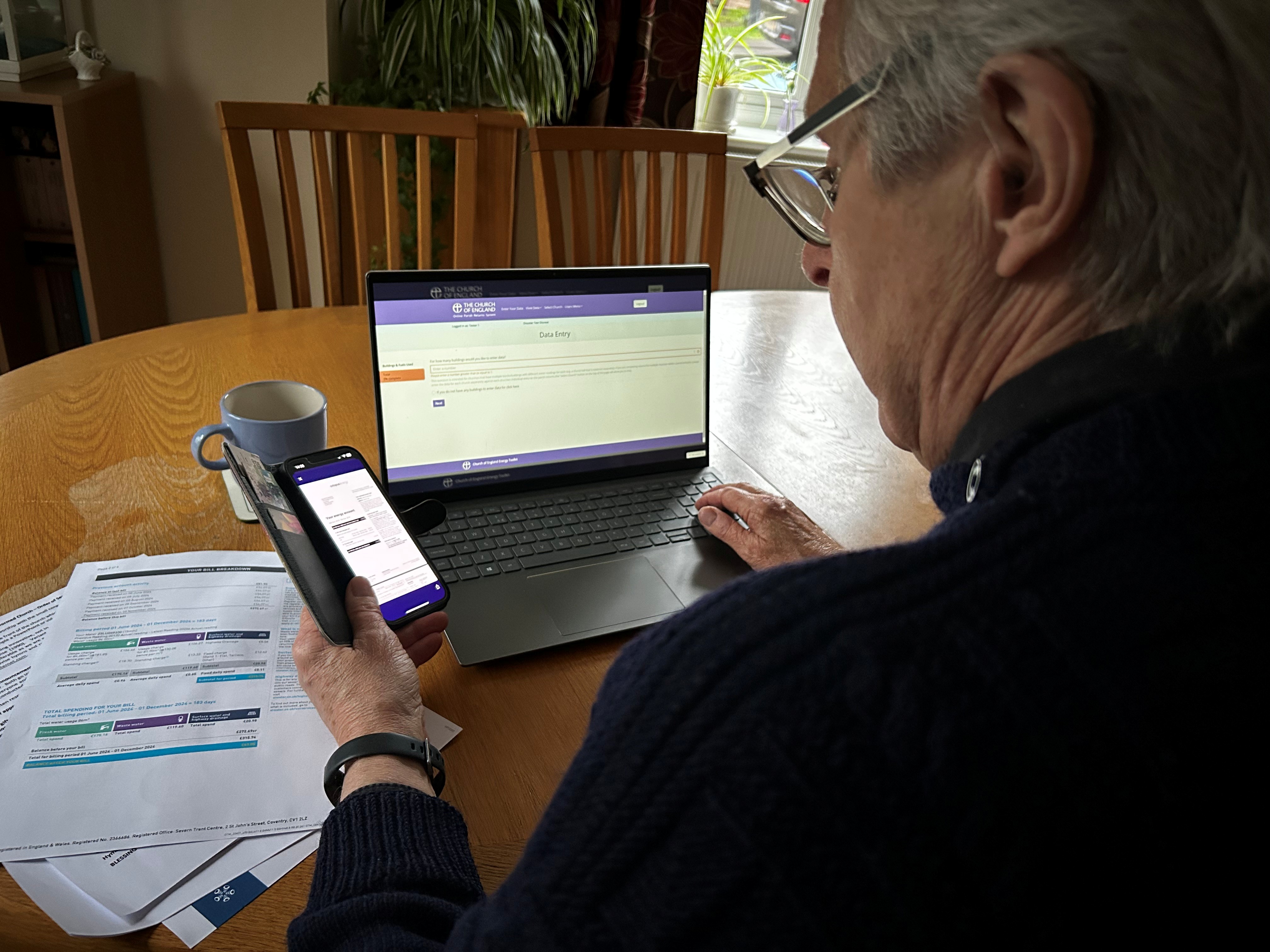 Man has phone and laptop open with paper bills to the side