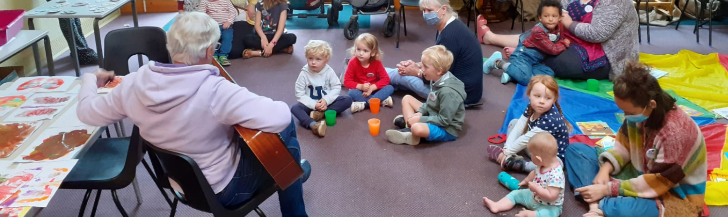 A group of children, parents and carers sit on the floor in and enjoy singing along to a guitar.