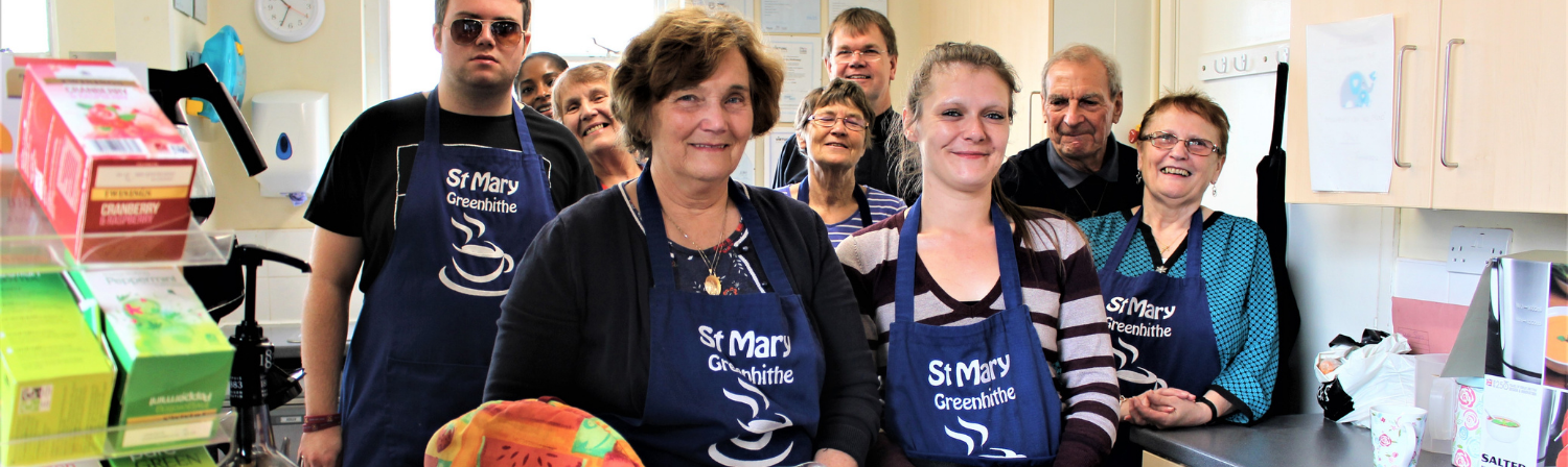 A mixed group of people stand smiling in a church hall kitchen as part of a church community cafe.