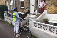 ...An Anna Chaplain stands with her bicycle and speaks from the bottom of some steps to an elderly lady standing in the doorway of her house.