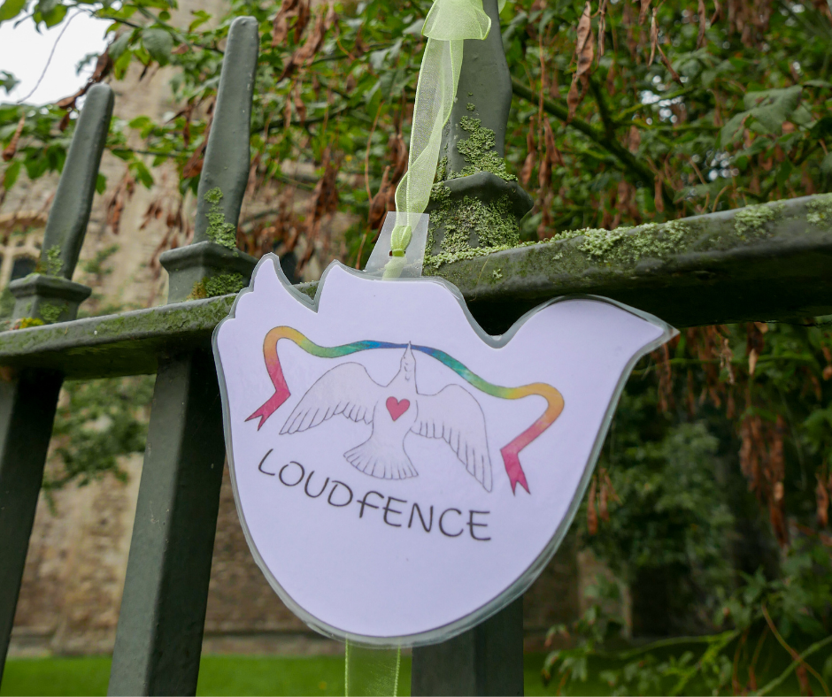Paper dove with LOUDfence image hangs from a yellow ribbon on railings around Rochester Cathedral