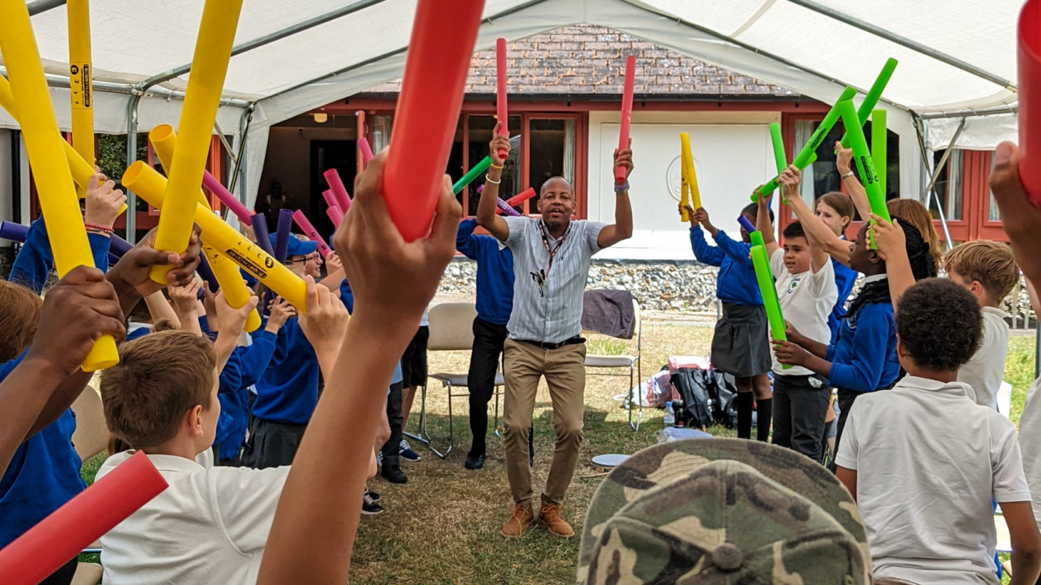 A large group of children gather around a volunteer as they hold colourful foam tubes in the air as part of a group activity.