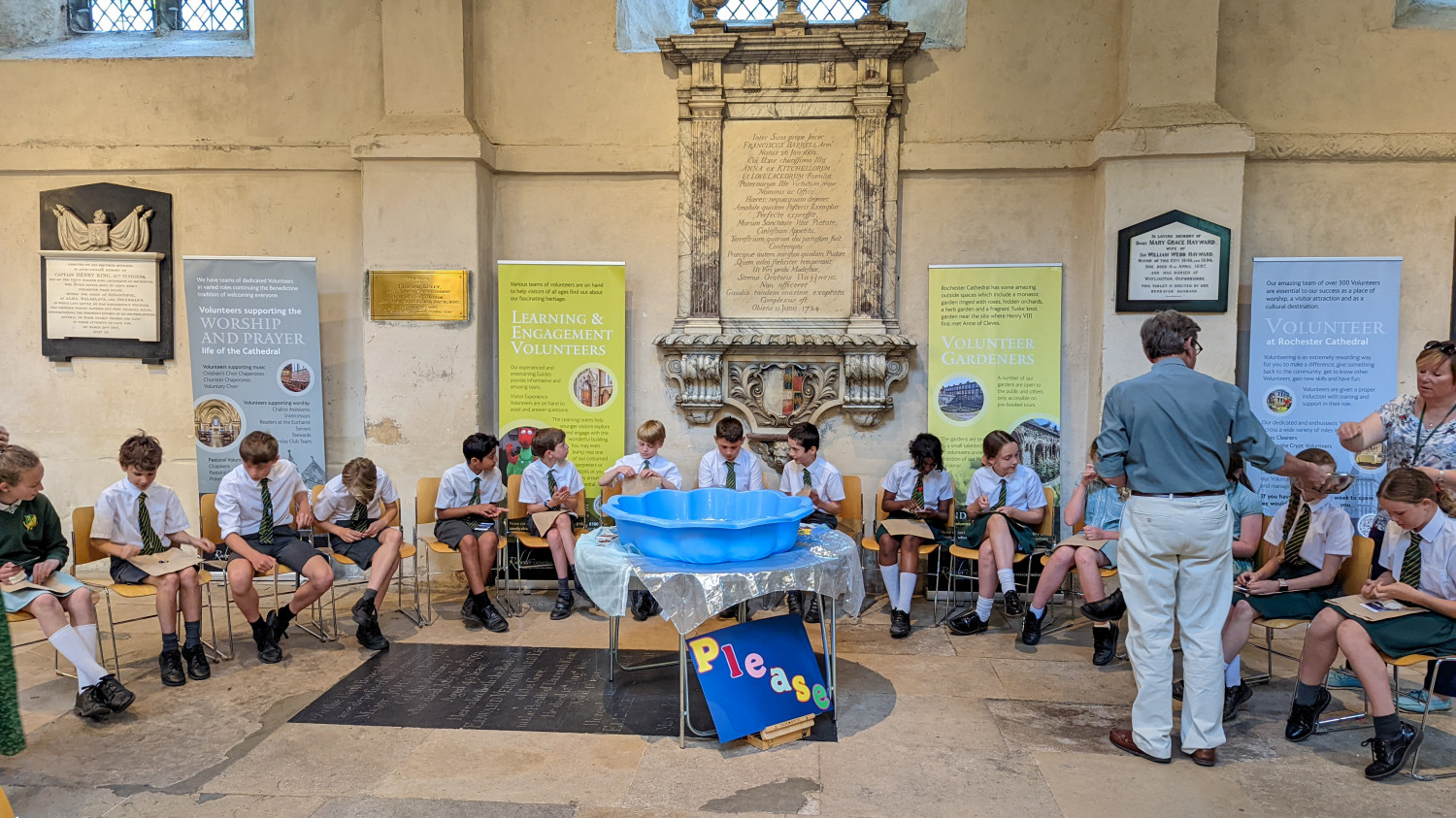 Children sit in a semi circle on chairs in the Cathedral. An empty blue sand pit is in the centre on a table. a colourful sign under it says the word, 'please'.