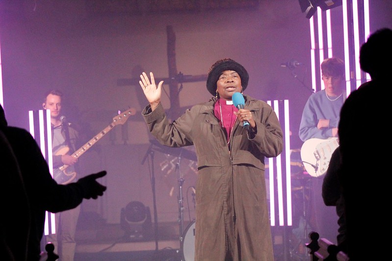 With a band in background, Bishop Rose speaks into a microphone.